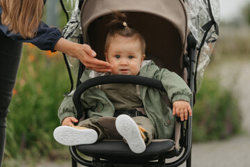 A female toddler is sitting in the stroller on a cloudy day. In a green village, a young serious girl in a raincoat is in a baby carriage. A mother is touching the cheek of her daughter.