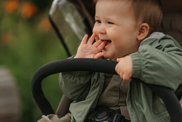 A close portrait of a happy female toddler who is putting her fingers in her mouth in the stroller on a cloudy day. In a green village, a young girl in a raincoat is in her baby carriage.