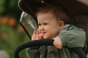 A close portrait of a female toddler who is putting her fingers in her mouth in the stroller on a cloudy day. In a green village, a young girl in a raincoat is in her baby carriage.