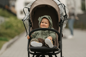 A happy female toddler is holding the safety bumper bar of the stroller on a cloudy day. A young...