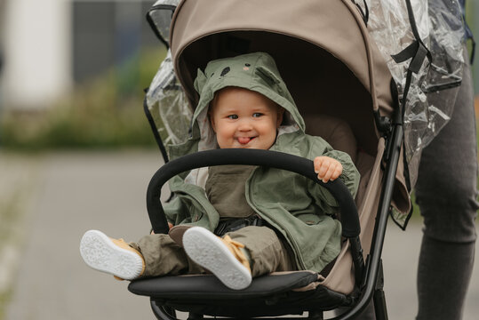 A female toddler with her tongue out is sitting in the stroller on a cloudy day. A young girl in a baby carriage in a village green.