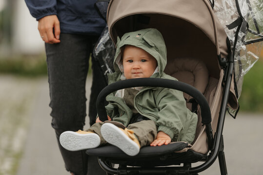 A female toddler is sitting in the stroller on a cloudy day. In a green village, a young girl in a raincoat is in a baby carriage. A mother is near her daughter.