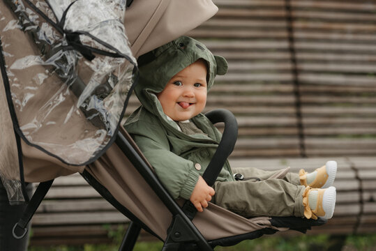 A photo from the side of a nice female toddler with her tongue out who is sitting in the stroller on a cloudy day. A young girl in a baby carriage in a village green.