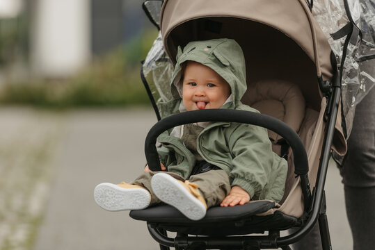 A happy female toddler with her tongue out is sitting in the stroller on a cloudy day. In a green village, a young girl in a raincoat is relaxing in a baby carriage.