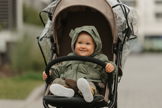 A female toddler is holding the safety bumper bar of the stroller on a cloudy day. A young girl in the hood of her raincoat is in her baby carriage in a park at noon.