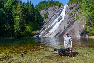 An adventurous athletic male hiker standing at the base of a waterfall with his Bernese Mountain...