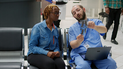Naklejka premium Male nurse showing bones radiography results to patient sitting in hospital reception lobby. Specialist and young woman looking at x ray scan to see healthcare diagnosis in waiting area.