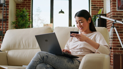 Fototapeta na wymiar Happy young adult woman inserting credit card credentials on on shopping site for online payment. Asian customer sitting on couch at home while paying for products on digital store.
