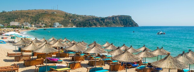 Landscape of beautiful clean sand and pebble beach with beach umbrellas and sun loungers on mountains background. Himare. Albania.