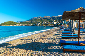 Landscape of beautiful clean sand and pebble beach with beach umbrellas and sun loungers on mountains background. Himare. Albania. - obrazy, fototapety, plakaty
