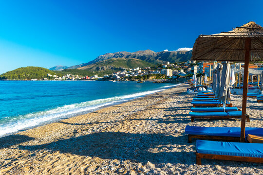 Landscape of beautiful clean sand and pebble beach with beach umbrellas and sun loungers on mountains background. Himare. Albania.