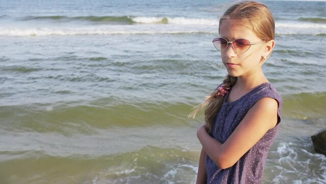 A girl in a blue dress with polka dots and sunglasses looks into the distance on the beach