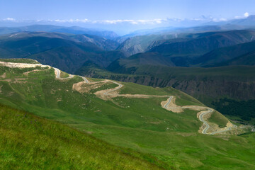 Naklejka premium winding mountain road with serpentines, green valley, high blue mountains, majestic nature, mountain landscape, travel, tourism, road adventure, direction, space for text, wallpaper, summer