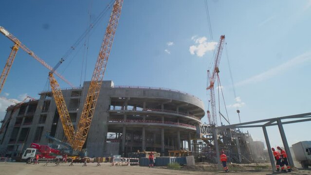 Powerful tower cranes and unfinished stadium building with workers at large construction site on sunny day. Sportive arena