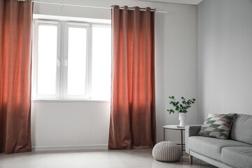 Vase with eucalyptus on table, sofa and pouf near red curtains in room