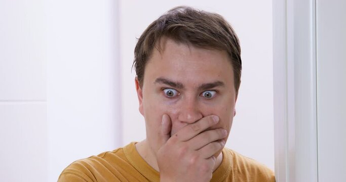 Plump man looks surprised to see mess standing at entrance in bedroom. Portrait of guy covering mouth with hand and moving eyes to sides closeup