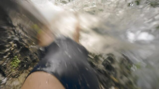 A girl in blue swimsuit touching powerful waterfall stream on tropical. Caucasian sexy girl stands under a rock from which a stream of mountain river flows on sunny day. Slow motion