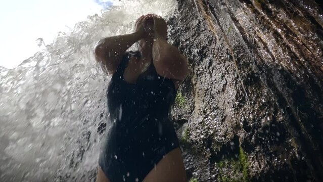A slender girl in a blue swimsuit stands under flow of a tropical waterfall waterfall. a sexy girl stands under a rock, from which a stream of a mountain river flows down on a sunny day. Slow motion