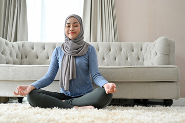 Peaceful millennial Asian Muslim woman practicing yoga, concentrated meditating