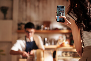 Young woman filming video of barista making coffee for social media account of coffeeshop, selective focus