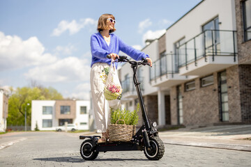 Happy stylish woman going home with fresh vegetables in mesh bag, driving on electrical scooter at residential district. Concept of modern eco-friendly lifestyle and sustainability © rh2010