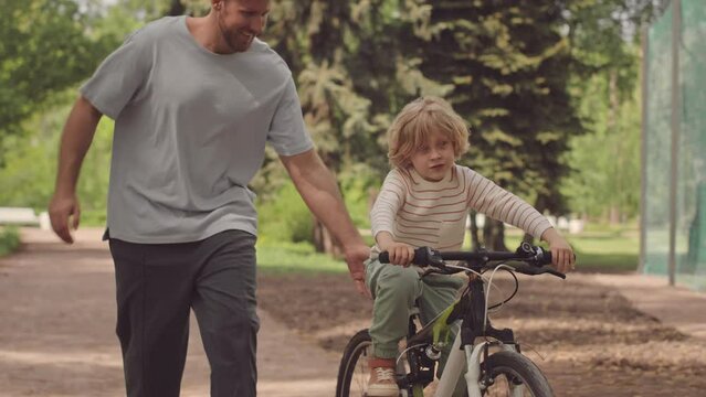 Slowmo of cheerful little boy riding bicycle along trail in park and his caring father supporting him, helping to keep balance