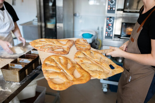 A woman in a bakery takes traditional French Fougasse bread out of the oven. Front view.