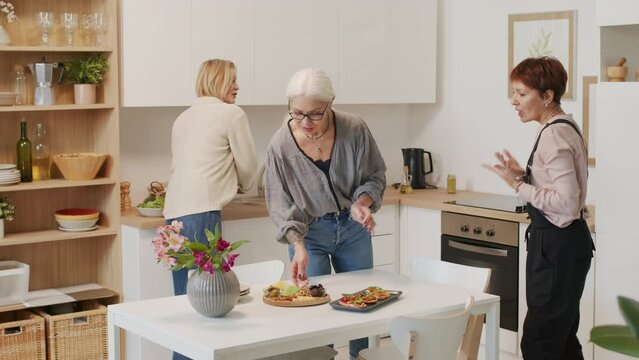 Three mature women serving kitchen table with starters and red wine in glasses, gathering together for sharing and discussing news