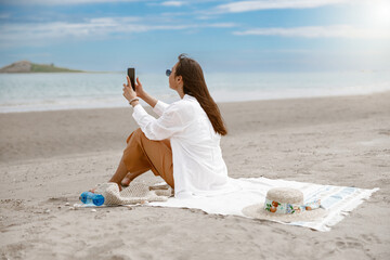 Beautiful woman in sunglasses sitting on beach towel near sea and using phone. Blurred background