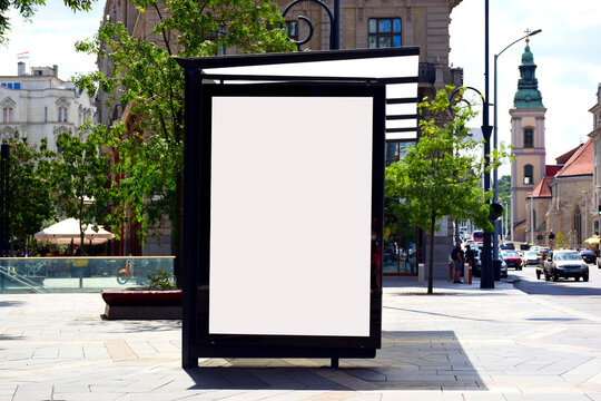 bus shelter at a busstop. blank billboard ad display. empty white lightbox sign. glass and aluminum frame structure.  city transit station. urban street setting. outdoor advertising. green trees.