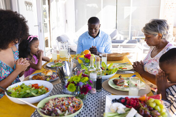 Multiracial multigeneration family with meal on table praying before having lunch at dining table - Powered by Adobe