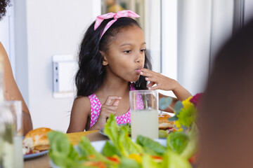 Biracial cute girl wearing pink headband licking fingers while having lunch at dining table at home - obrazy, fototapety, plakaty