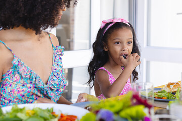 Biracial girl eating food and looking away while sitting with mother at dining table for lunch
