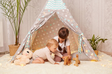 Portrait of dark haired little girl with braids playing with wooden toys in the wigwam together with her baby sister, sitting on floor on soft carpet, sibling spending time together. © sementsova321