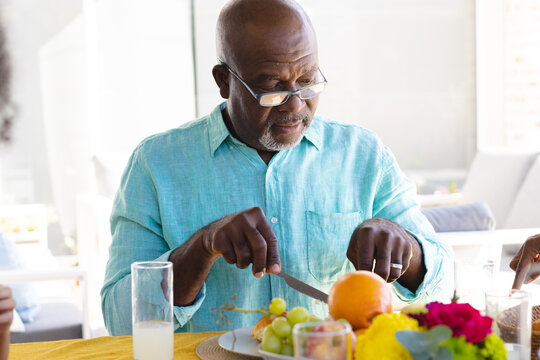 African american bald senior man wearing eyeglasses eating fruits for lunch on dining table at home