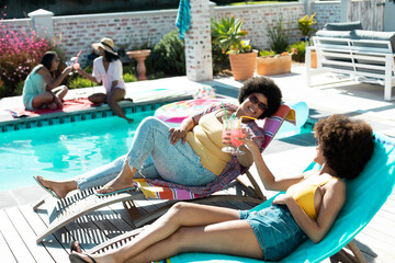 High angle view of biracial females toasting drinks while enjoying relaxing at poolside in summer - Powered by Adobe