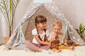 Portrait of little girl with pigtails wearing white t shirt playing with wooden toys in the wigwam together with her baby sister, elder kid looking after infant child, having fan in teepee tent. © sementsova321
