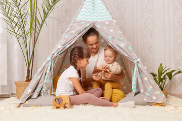 Indoor shot of mother with two kids sitting together in wigwam teepee at home, mom playing with preschool and infant children, showing wooden eco toy to her baby. © sementsova321