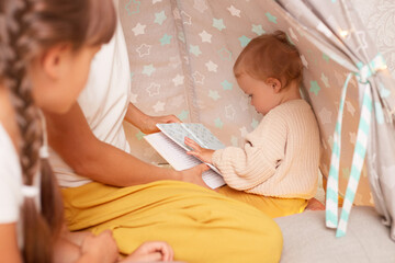 Closeup portrait of infant baby posing in wigwam with her sister with braids and faceless mother sitting near her, charming kid holding book in hands, playing with her family in teepee tent  © sementsova321