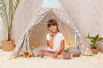 Indoor shot of little girl with pigtails wearing white t shirt playing in wigwam alone, sitting on the floor and talking via with telephone with her friend, looking at camera. © sementsova321