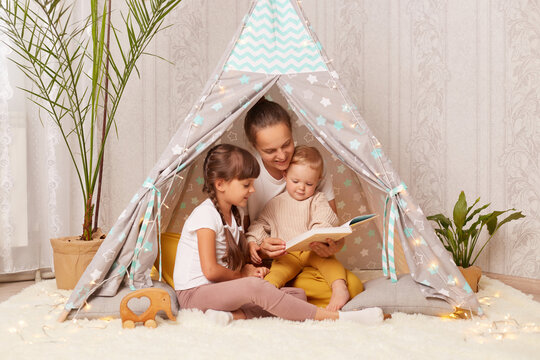 Image of woman with her little daughters reading book in hovel at home, Caucasian female sitting on floor in wigwam, read fairy tale to her kids, spending happy time together.