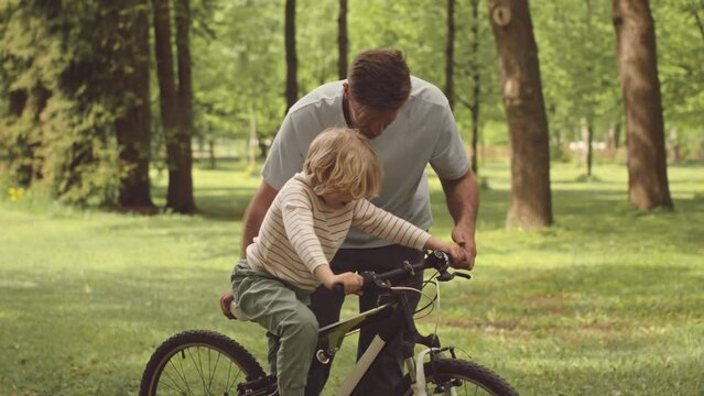 Slowmo of father teaching his 7 year old son to ride bicycle outdoors in park on sunny day