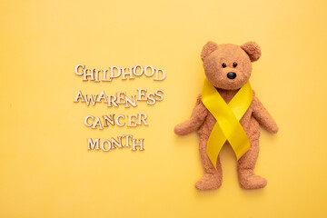 Childhood cancer awareness month text from wooden letters and teddy bear with yellow ribbon on yellow background
