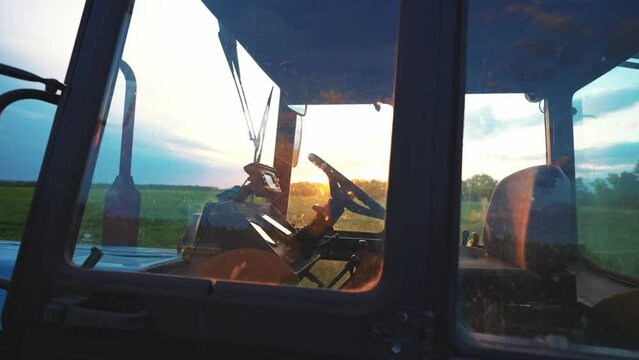Side view through window of tractor driver cab with steering. Car is standing on field against sunset. Management agriculture with help of equipment. Simplification of production process on farm.