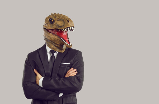 Funny Man With Dinosaur Mask On Head With Arms Crossed Symbolizing Aggressive And Assertive Strategy To Achieve Goals In Career Or Business Dressed In Formal Suit Stands On Studio Background