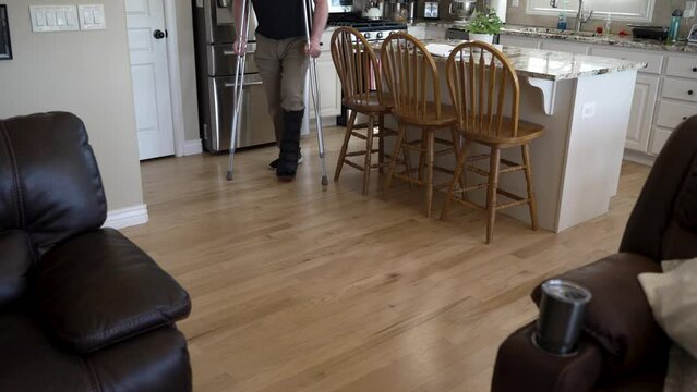Man with a broken leg in an orthopedic boot uses crutches to get around his home - tilt down