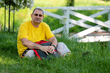Naklejka na ściany i meble a senior man with a backpack sits on the grass in a park on a sunny day