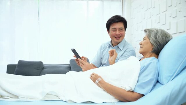 Son of an elderly woman patient using tablet to make video calls with family. Female patient lying in bed receiving saline solution greet the camera with a smiling and encouraging face.