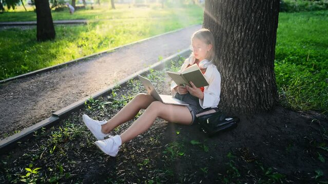 Beautiful teenage girl sitting in park with laptop and reading book aloud. Young female student is preparing for session and exams on outdoors. Leisure activity on back of summer landscape of nature.