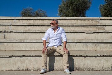 handsome mature man with beard and grey hair is sitting on the steps of an open-air auditorium. The man looks at different places. Senior concept, travel and tourism.